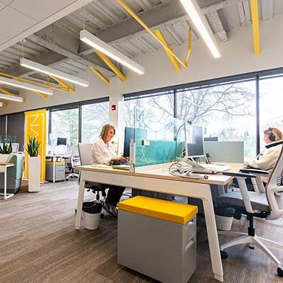 Two members at white sit-stand desks below HAYVN's yellow painted ceiling pipework